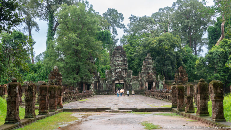 Wide stone approach lined with guardian figures leading to a temple gateway among tall trees.