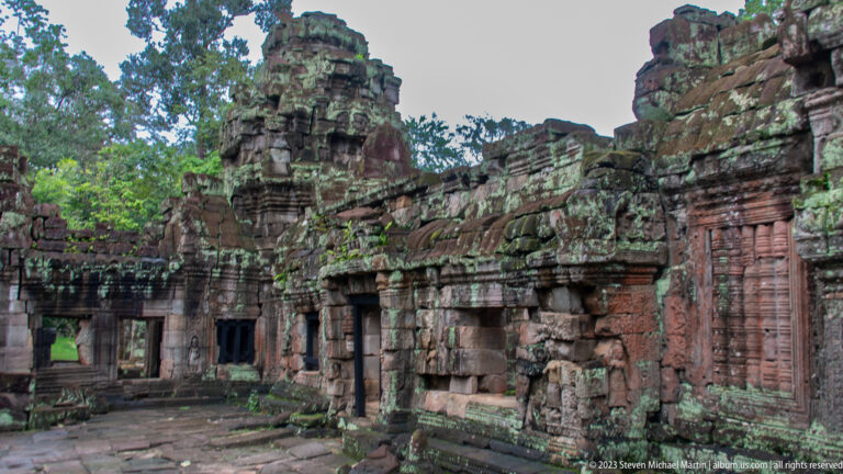 Oblique courtyard view of a long ruined sandstone wall with doorways, carved panels, and a tower.