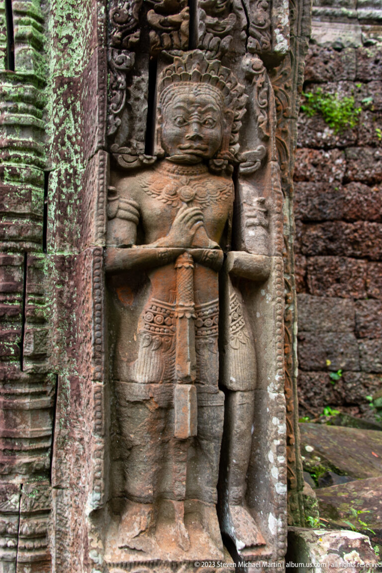 Large standing sandstone figure with crossed forearms, elaborate headdress, and long central ornament.