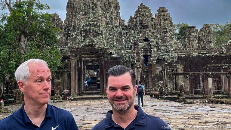 Two male visitors posing before a large towered sandstone gateway with people walking through it.