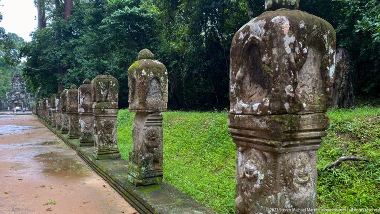Row of decorated sandstone posts receding along a temple path beside grass and forest.