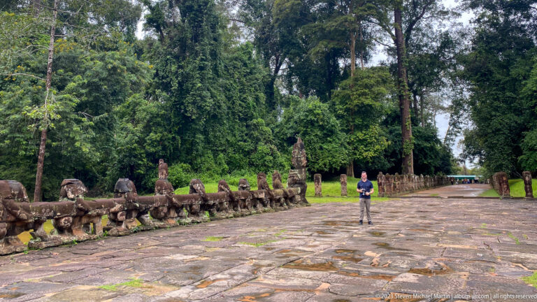 Wide stone causeway through forest with rows of guardian figures and a visitor standing near the center.