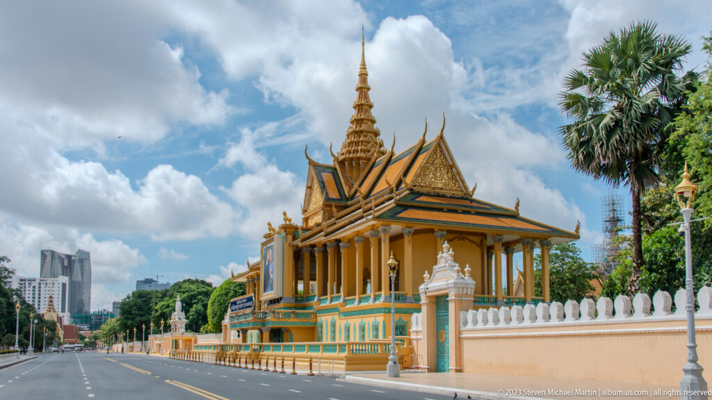 Royal Palace pavilion with gilded tiered roof and spire behind a long wall beside an empty Phnom Penh street.