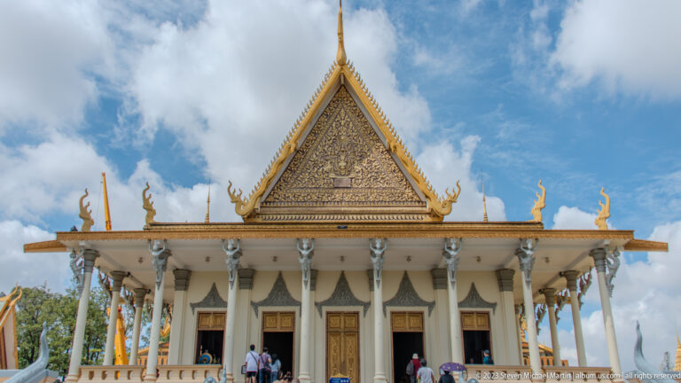 White palace pavilion with gold relief pediment, slender columns, dark doorways, roof finials, and visitors.