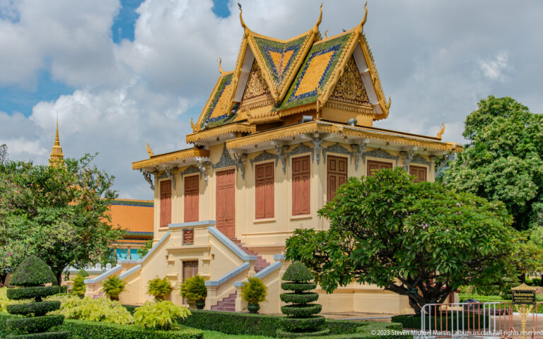 Two-level palace pavilion with red shutters, ornate Khmer roof, twin stairways, and clipped garden trees.