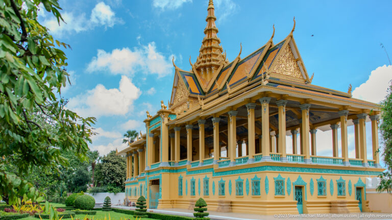 Large palace pavilion with tall cream columns, turquoise and gold base, layered roof, central spire, and garden shrubs.
