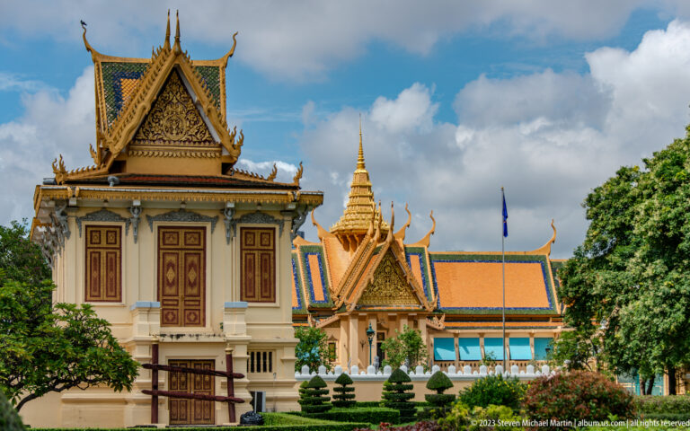 Small shuttered palace pavilion in front of trees, clipped shrubs, gilded gables, and a distant spire.