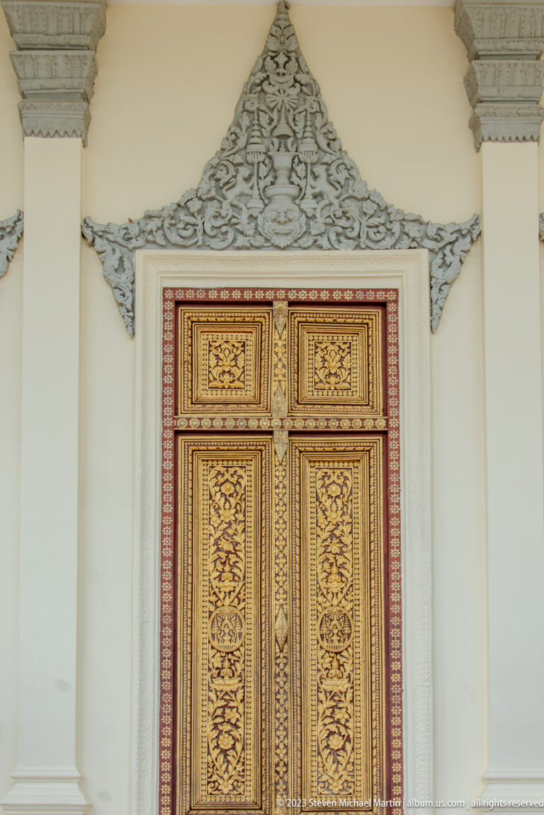 Red and gold carved double doors beneath an elaborate silver-toned ornamental crest on a pale wall.