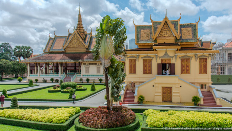 Formal palace garden with geometric hedges and circular beds leading toward a small elevated pavilion and larger buildings.