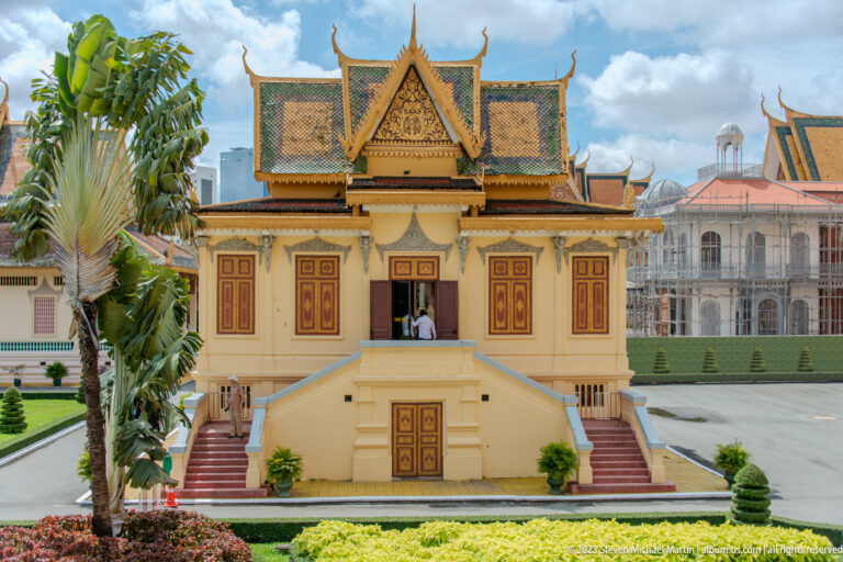 Symmetrical cream pavilion with red-and-gold shutters, twin stairways, ornate roof, and clipped garden beds.