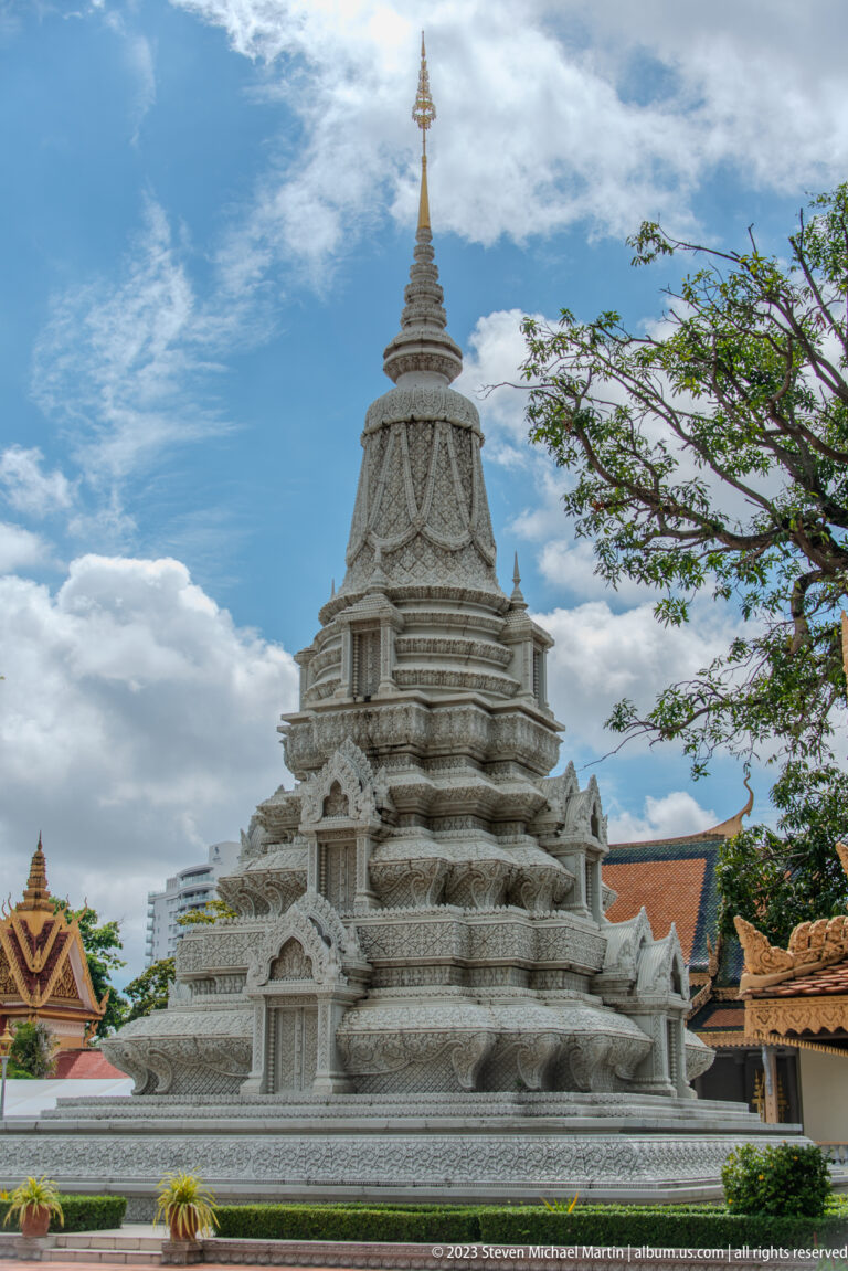 Large white carved stupa with stepped tiers, dense relief ornament, and a gilded finial under a blue sky.