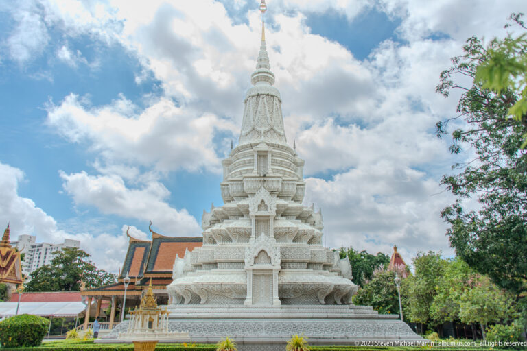 White tiered stupa with carved niches, repeating cornices, and a slender finial beside palace roofs.