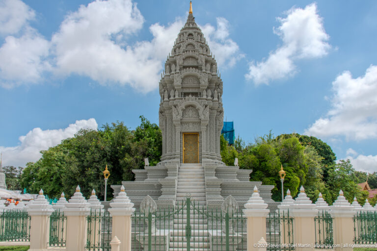 Tall white memorial stupa with carved façade, gilded door, narrow stair, smaller stupas, trees, and green fence.