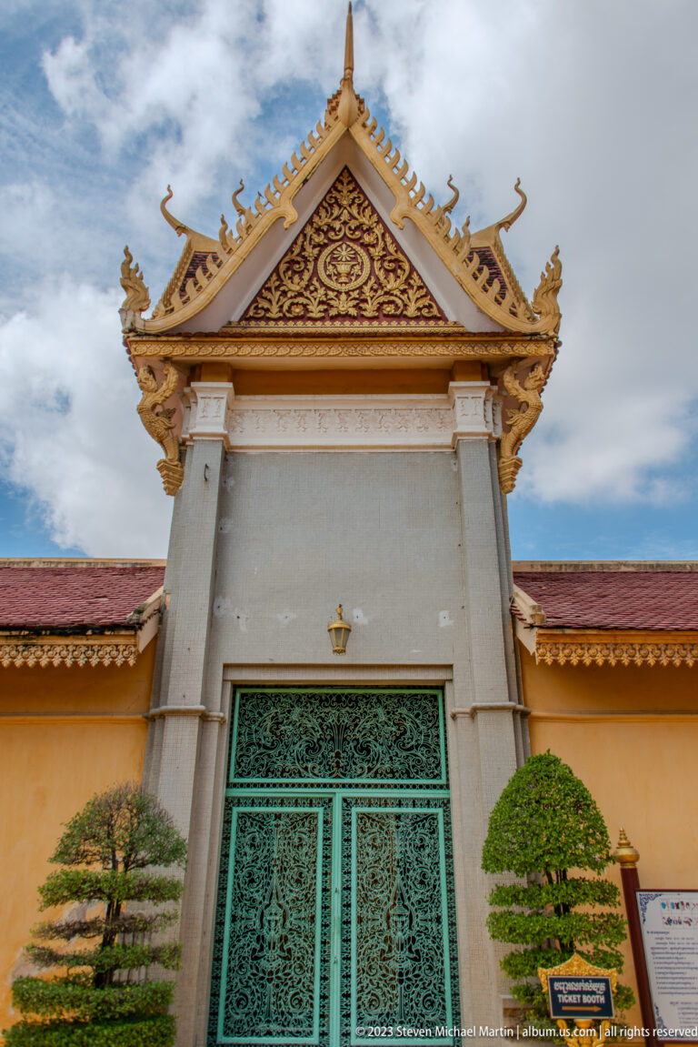 White palace gateway with gilded triangular pediment, turquoise doors, clipped trees, and a red roof.