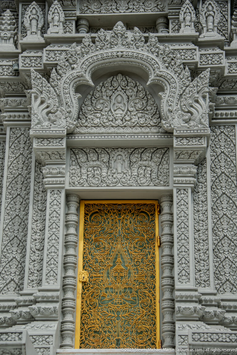 Golden ornamental door framed by dense white floral relief, pilasters, niches, and an arched crest.