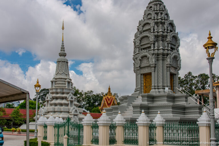 Two white memorial stupas, one broad and tiered and one tall with a gilded door, behind a green fence.