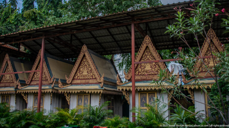 Traditional wooden palace building with repeated ornate triangular gables beneath a large protective canopy and surrounded by plants.