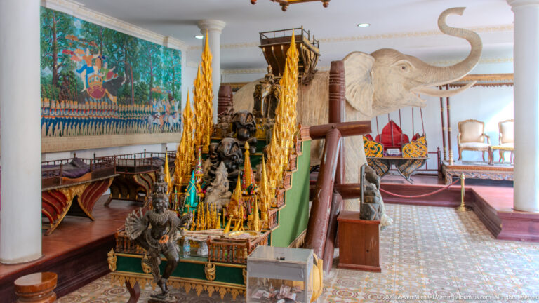 Palace interior display with gilded ceremonial objects, decorated furniture, rope barriers, and a large white elephant sculpture.