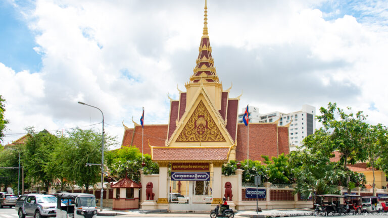 National Museum of Cambodia entrance with tall spire, ornate gable, red walls, flags, guardians, cars, and motorbikes.