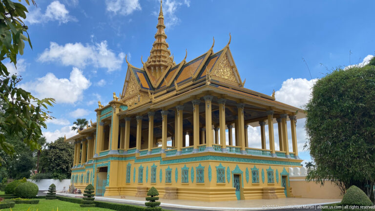 Oblique palace pavilion with tall columns, turquoise-and-gold base, layered roof, central spire, lawn, and clipped shrubs.