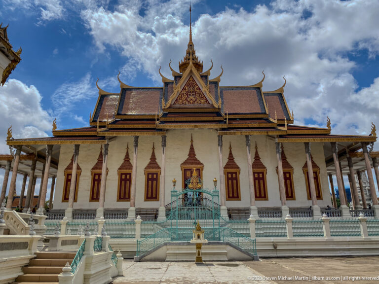 Centered front view of the Silver Pagoda with white columns, red doors, turquoise stairs, ornate roof, and cloudy sky.