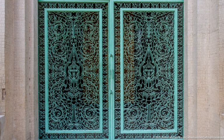 Close view of turquoise metal palace doors covered with symmetrical scrollwork and pointed decorative motifs.