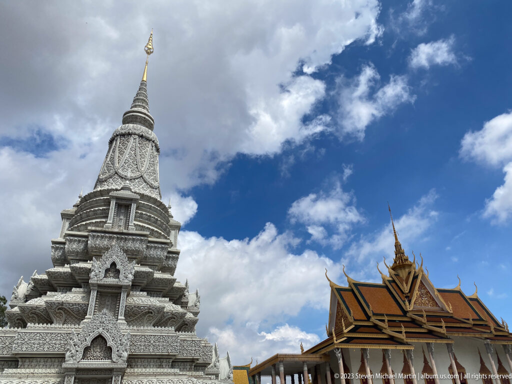 Low-angle view of a white carved stupa beside a gilded palace roof against blue sky and clouds.