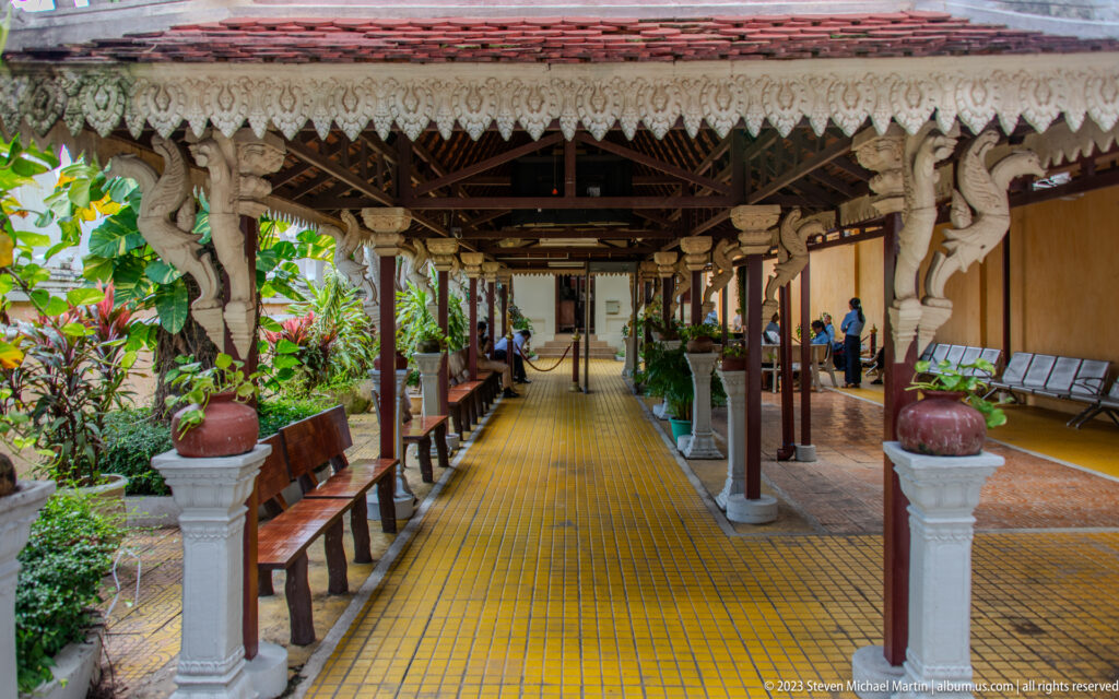 Covered palace corridor with yellow tiles, carved brackets, dark columns, benches, plants, and visitors.