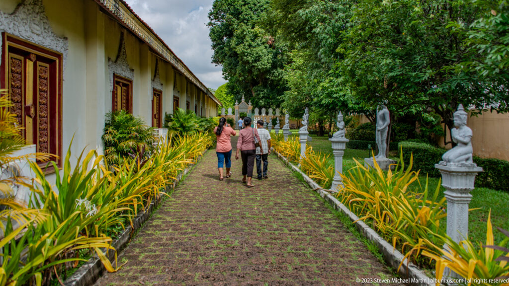 Visitors walking on a brick garden path beside palace buildings, yellow-green plants, trees, and white decorative figures.