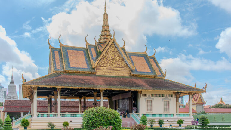 Open-sided palace pavilion with ornate tiered roof, cream walls, turquoise trim, lawn, and clipped shrubs.