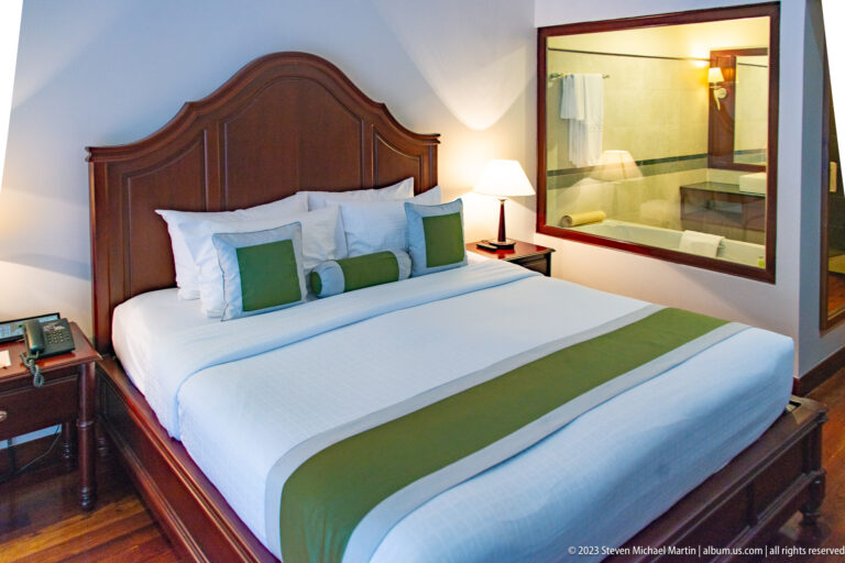 Hotel room with dark wooden bed, green-and-white linens, bedside lamps, and a large window into the bathroom.