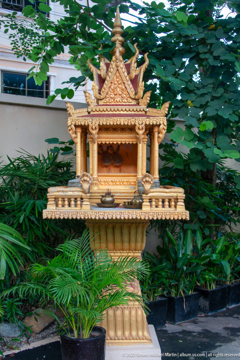 Golden spirit house with tiered roof and pointed finials standing on a pedestal among tropical plants.