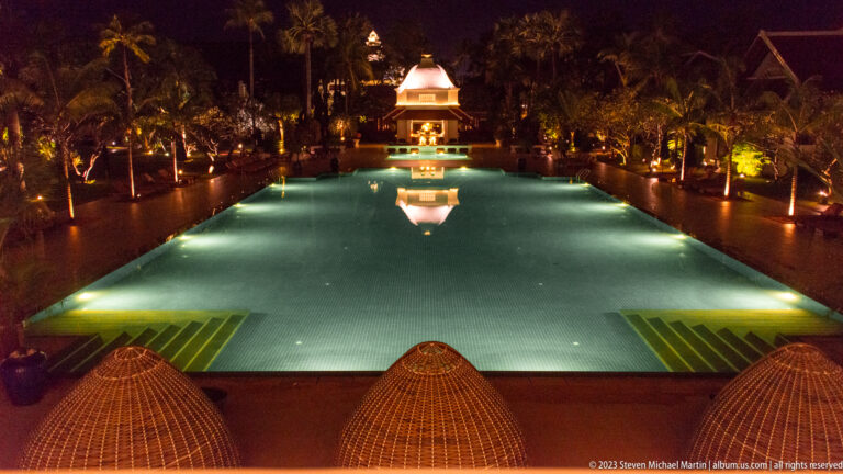 Long illuminated hotel pool leading toward a small lit pavilion, with palms and garden lights at night.