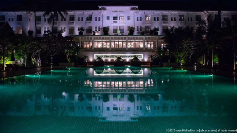 Grand Hotel d’Angkor illuminated at night and reflected across a dark turquoise swimming pool.