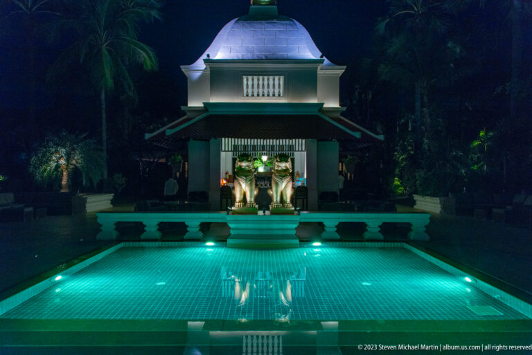 Small domed pavilion illuminated beyond a turquoise pool and reflected in the water at night.