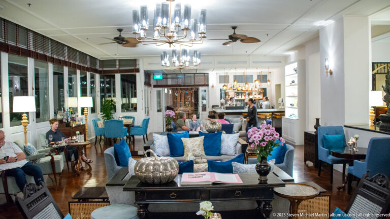 Bright hotel lounge with blue chairs, chandeliers, flowers, dark tables, guests, and a bar.