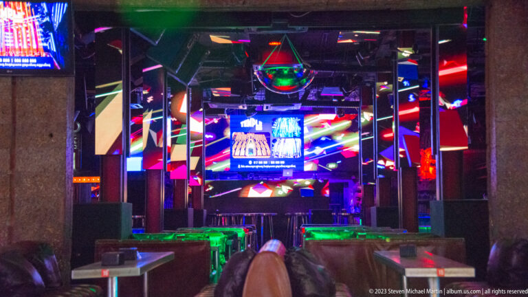 Neon-lit nightclub interior with mirrored columns, screens, empty tables, stools, and a bright stage area.