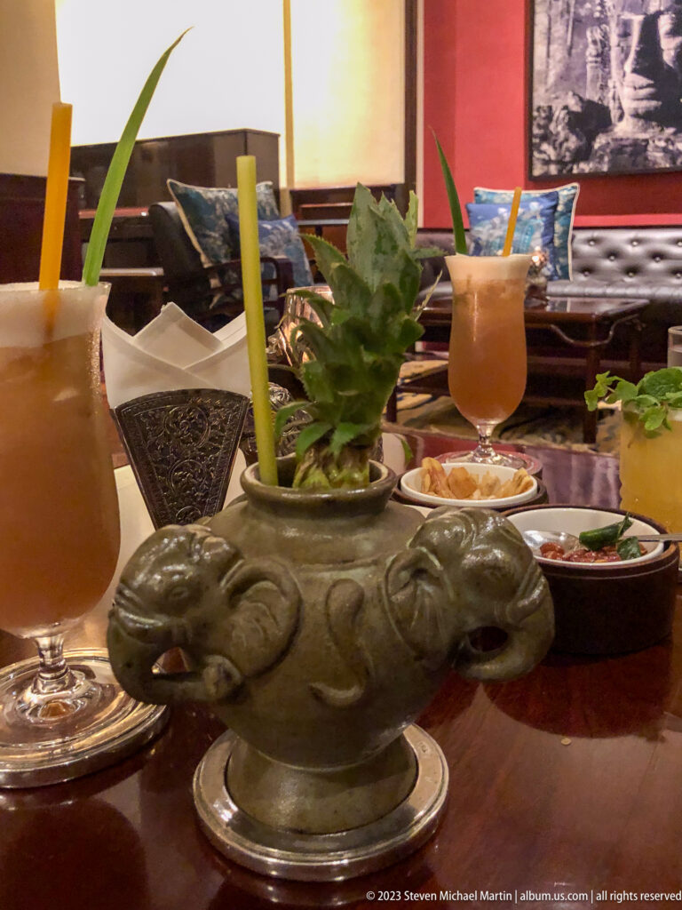 Elephant-shaped cocktail vessel with pineapple garnish beside a tall drink and small dishes.