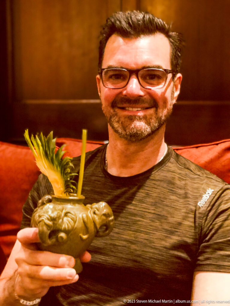 Smiling man holding an elephant-shaped cocktail vessel with pineapple garnish.