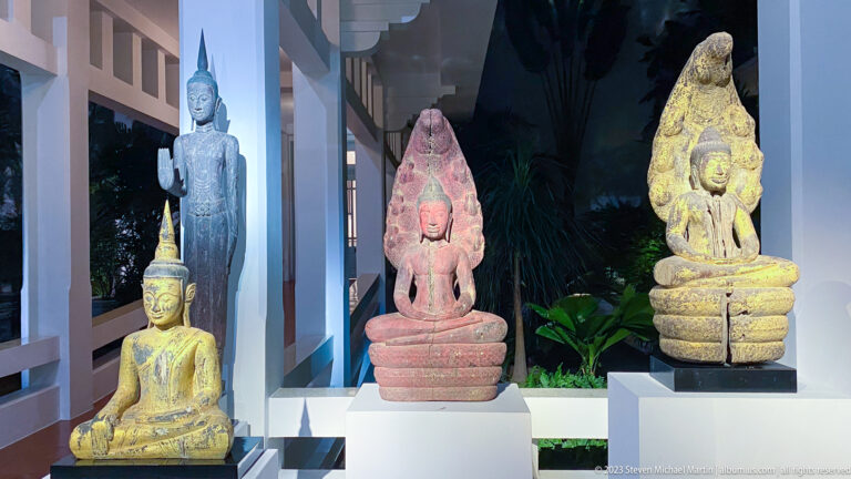 Several Buddha sculptures on white plinths, including seated and standing figures and naga-protected images.