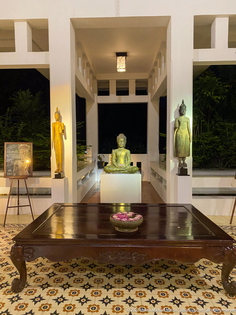Open white pavilion with seated Buddha at center, flanking sculptures, low table, and patterned floor.