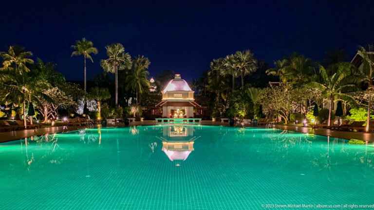 Long turquoise hotel pool leading to an illuminated pavilion amid palms and tropical gardens at night.