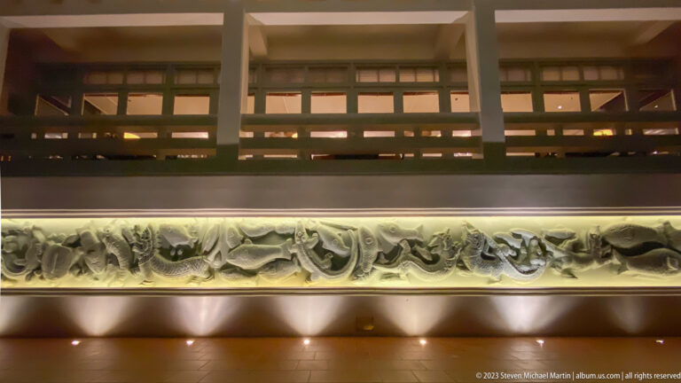 Long illuminated relief of intertwined aquatic or serpent-like creatures beneath a hotel balcony.