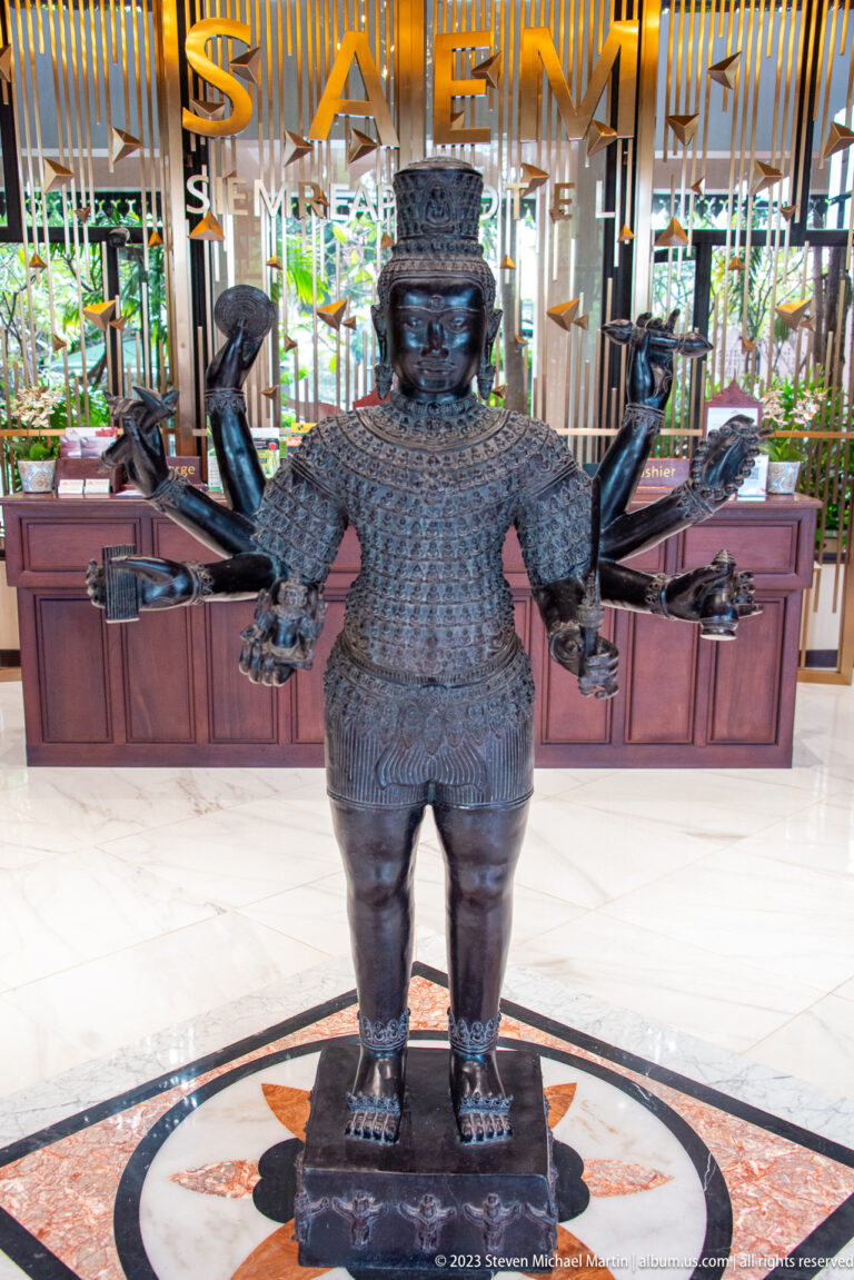 Dark four-armed sculptural figure with headdress and jewelry standing on a circular floor medallion in a hotel lobby.
