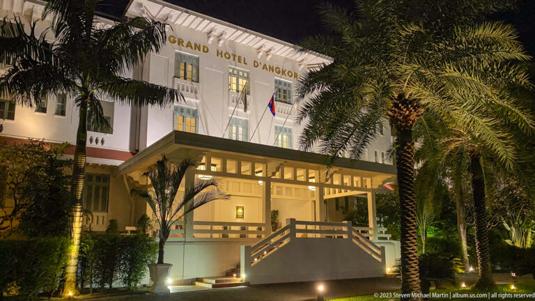 Grand Hotel d’Angkor entrance illuminated at night behind tall palms and landscaped grounds.