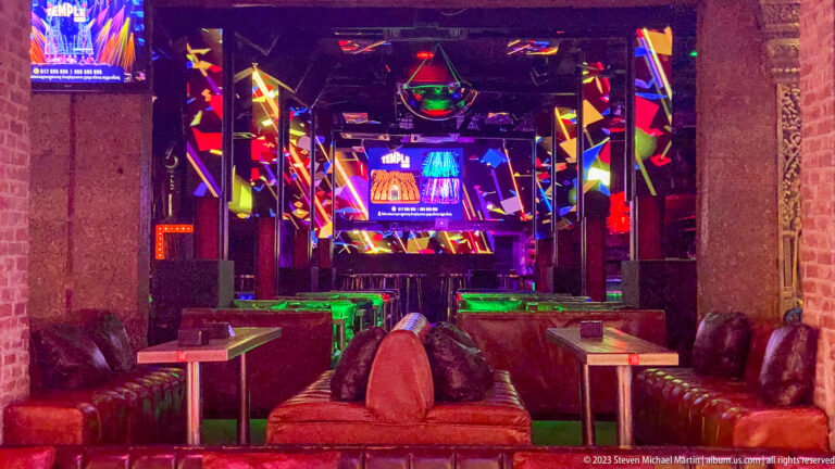 Modern neon-lit bar interior with mirrored columns, lounge seating, and colorful lighting.