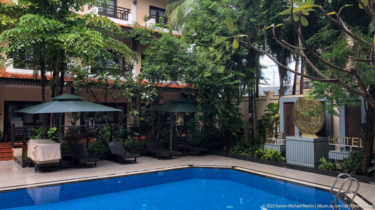 Hotel swimming pool surrounded by tropical gardens, trees, and guest accommodations.