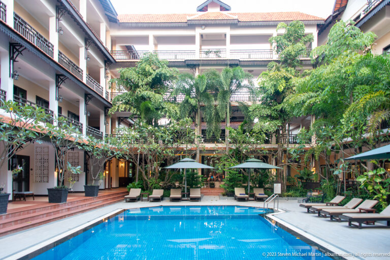 Blue swimming pool enclosed by multi-story hotel balconies, shaded walkways, lounge chairs, and tropical trees.