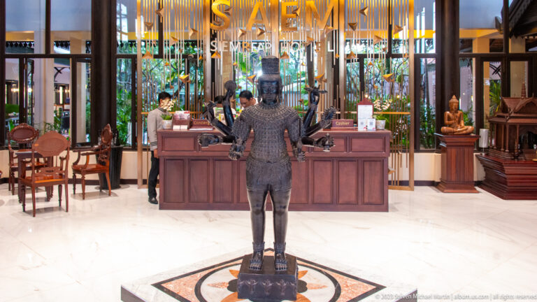 Rear view of a four-armed sculpture facing a wooden hotel reception desk beneath a suspended gold-toned screen.