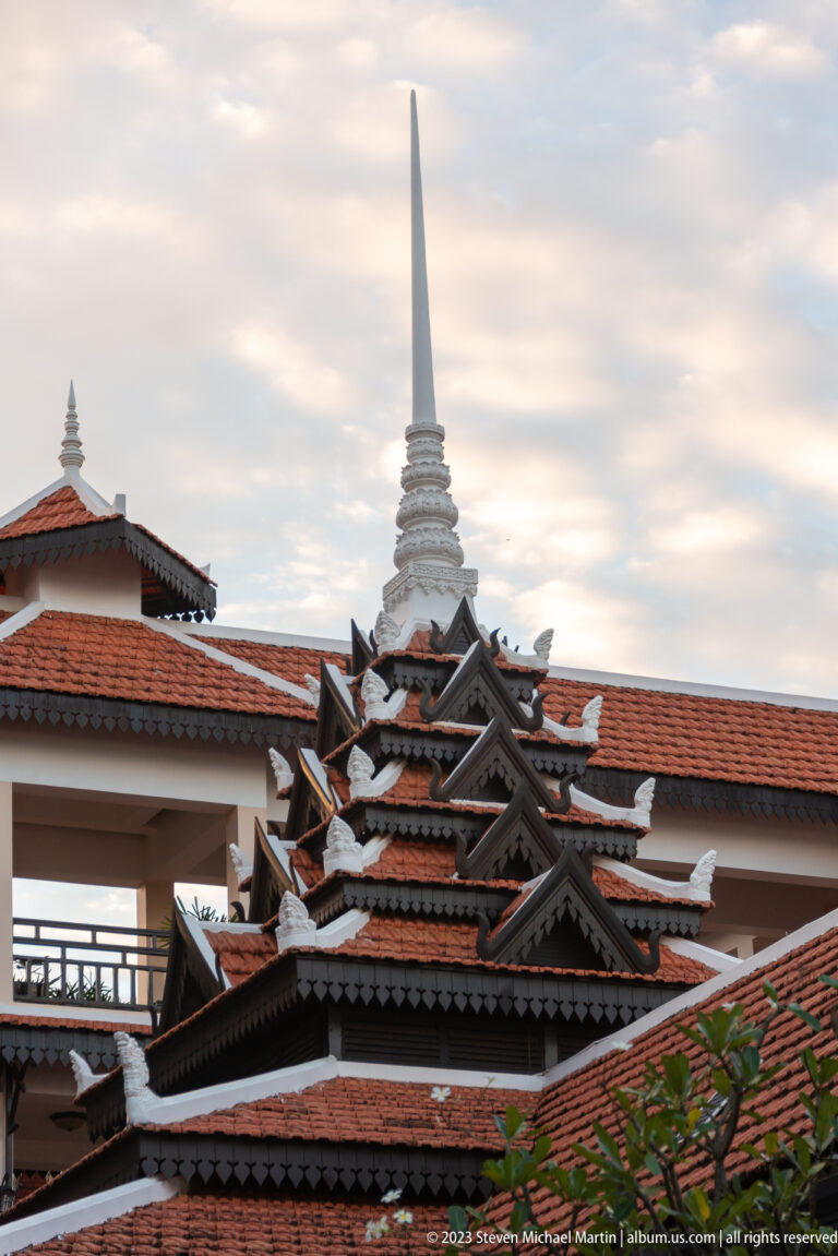 Layered dark gables and red tile hotel roofs silhouetted beneath a slender white spire and cloudy sky.